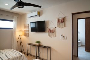 Modern bedroom with wall-mounted TV, decorative wall hangings, and minimalist furniture in a bright,.